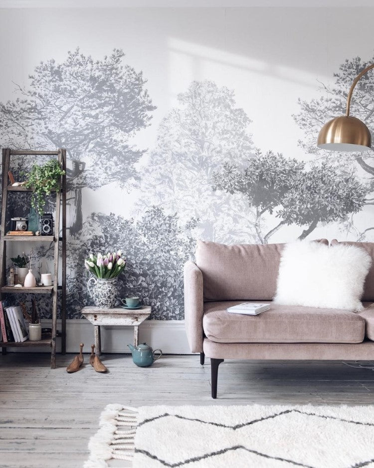 Classic Mua, Mural Wallpaper in Grey sets a cozy and elegant living room with a detailed tree and foliage design, a beige sofa with a white fluffy pillow, a small round table with a vase of fresh tulips, a shelving unit with books and plants, and a modern pendant light