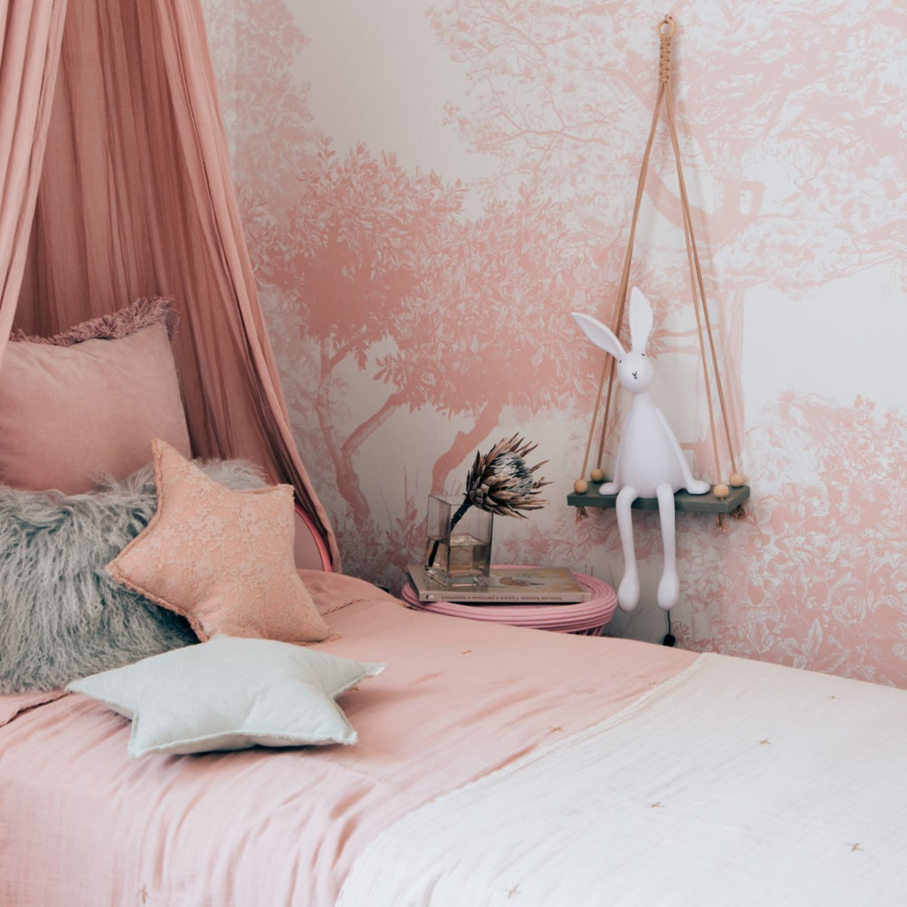 Classic Mua, Mural Wallpaper in Pink sets a cozy corner of a bedroom, a bed with pink bedding and various pillows, a long, flowing pink canopy, and a small wooden shelf with a white figurine of a rabbit, stacked books, and a vase with dried flowers.