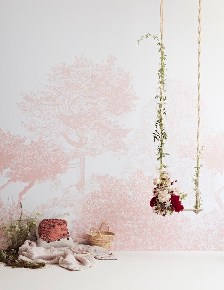 “Classic Mua, Mural Wallpaper in Pink adorns a wall, depicting a tranquil forest. A hanging floral arrangement adds a touch of nature, while a cozy seating area with cushions and a woven basket invites relaxation. The scene exudes calm and peace.