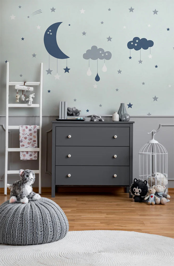 A child’s room under a Cloudy Night, Wallpaper, featuring a grey dresser, a white ladder shelf filled with books and toys, a knitted pouf, and plush toys on a patterned rug over wooden flooring.