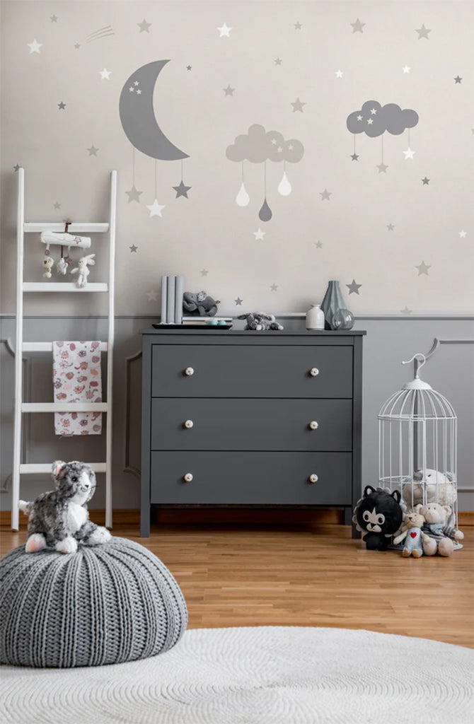 A children’s room featuring Cloudy Night, Wallpaper in Sand. Modern furniture includes a ladder-style shelf, a grey dresser, and a knitted pouf. A plush dog toy rests on the pouf. The room exudes a cozy and serene atmosphere.