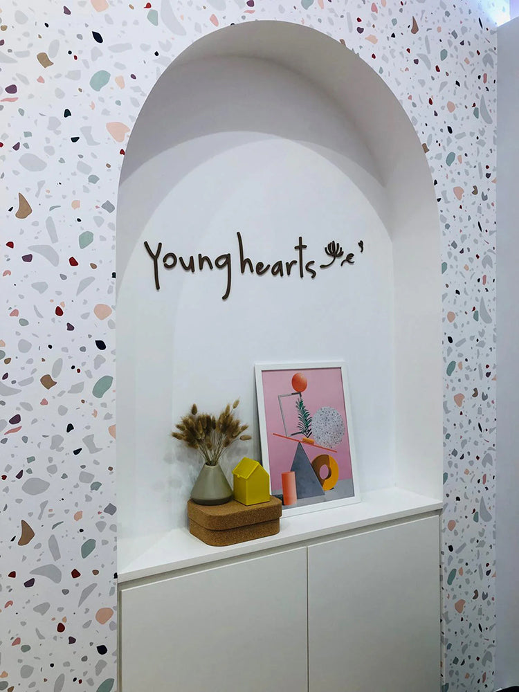Interior space with Confetti Terrazzo, Pattern Wallpaper, featuring multicolored speckles. Includes two arched doorways, one with a pink door, a wall sconce, and a ‘Young hearts’ text above a decorative shelf with a vase and framed artwork.