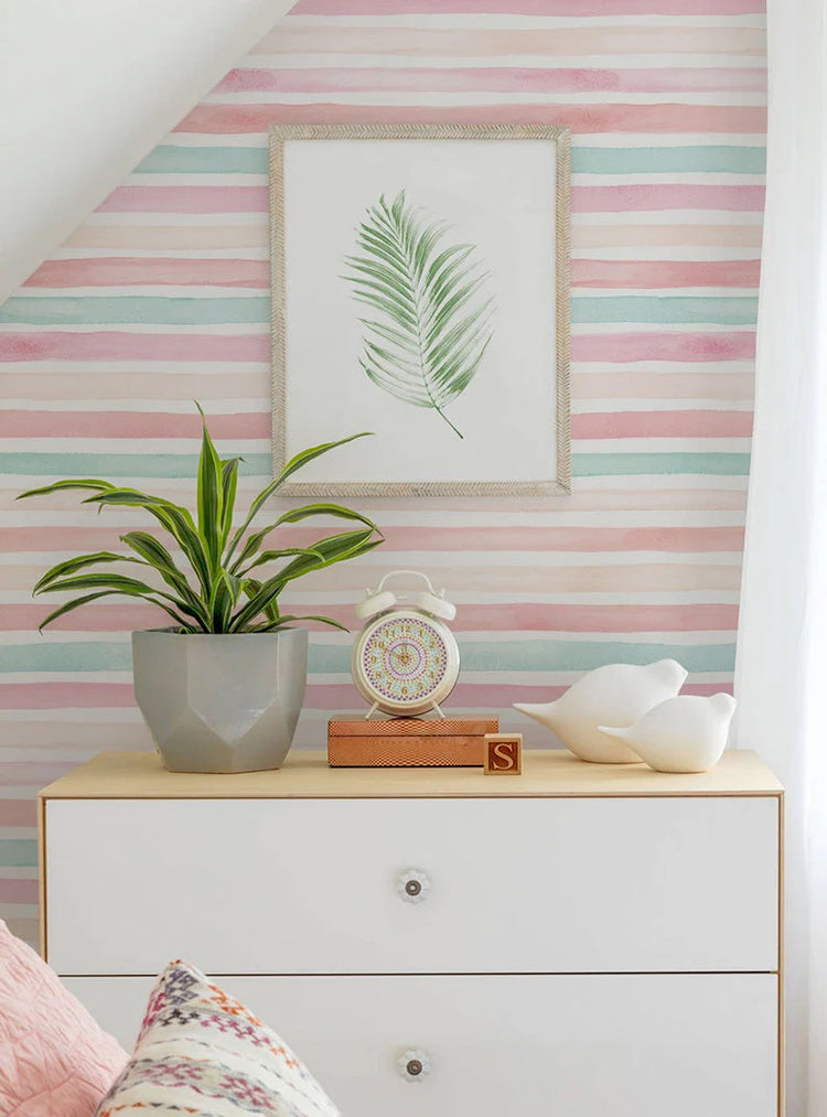 A tastefully decorated corner of a room featuring Coral Mint Stripes pattern wallpaper. A white dresser holds a plant, books, and decor, under a framed leaf art. The room exudes a sense of tranquility and elegance.