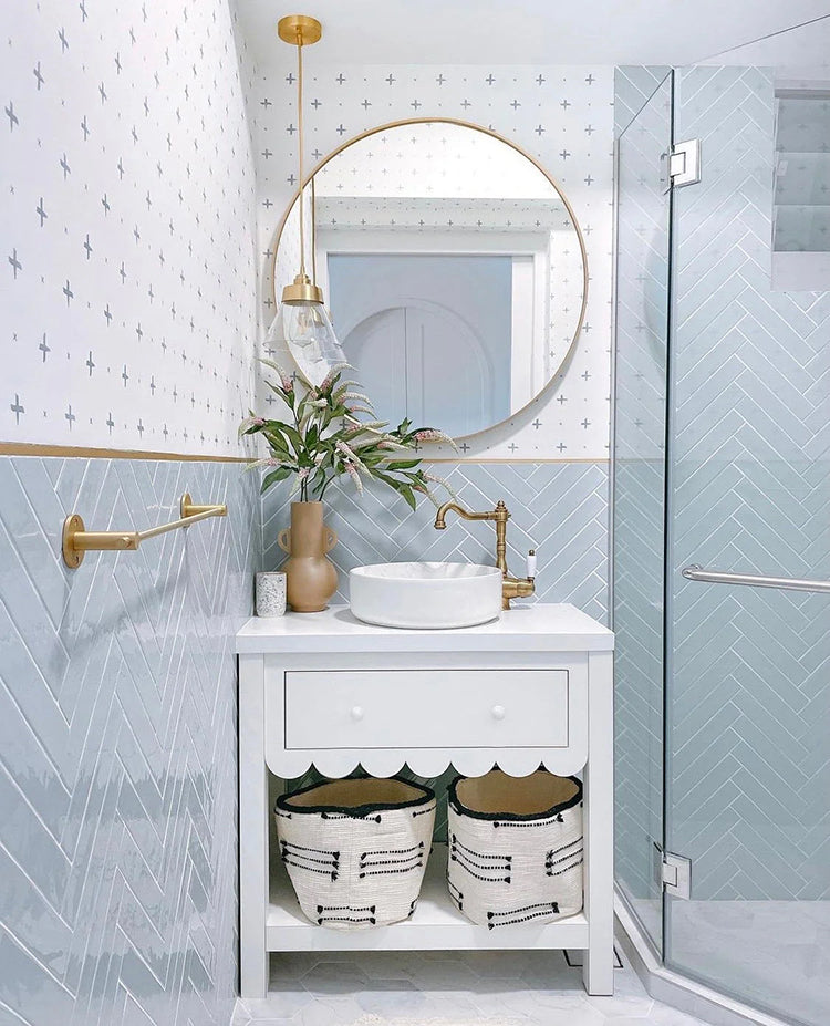 A bright, elegant bathroom with Cross Stitch, Blue Pattern Wallpaper. Features a white vanity with a round mirror, gold fixtures, a glass shower door revealing Cross Stitch, Blue Pattern Wallpaper, and decorative plants.