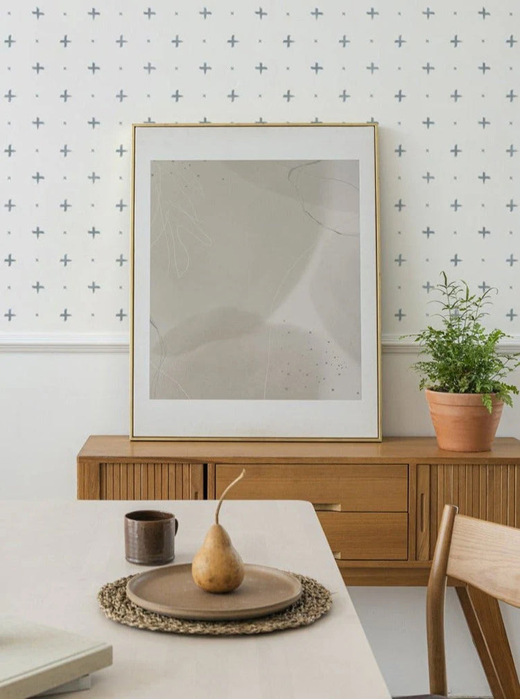 A modern room featuring Cross Stitch, Blue Pattern Wallpaper. The room includes a wooden sideboard displaying abstract art and a potted plant. A dining setup with a pear on a plate is also visible.