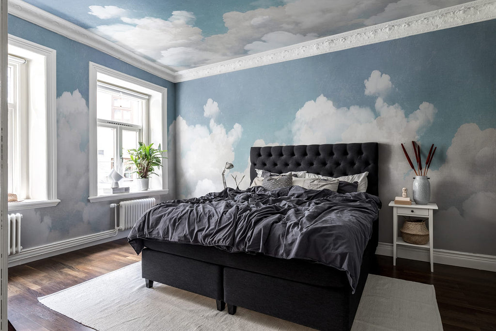 Bedroom with Cuddle Clouds, Mural Wallpaper in Blue, featuring a serene blue sky and fluffy white clouds on the upper walls and ceiling. The room includes a dark upholstered bed with gray bedding, a window, and a nightstand.