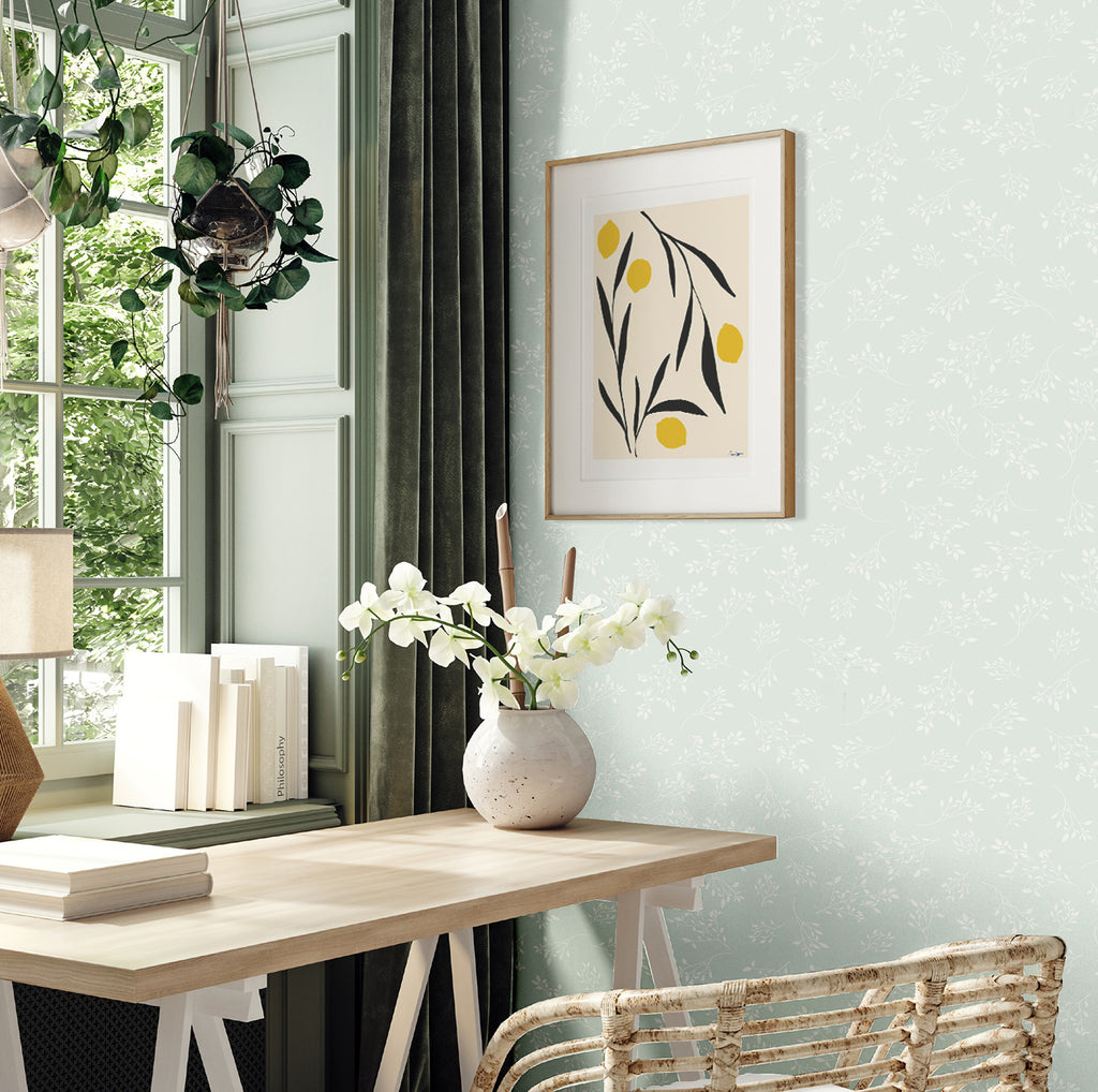 In an elegant interior, natural light filters through large windows, illuminating the subtle Delicate Foliages, Pattern Wallpaper in Blue. The room boasts a wooden desk, a collection of books, and a rattan chair, creating an inviting atmosphere.