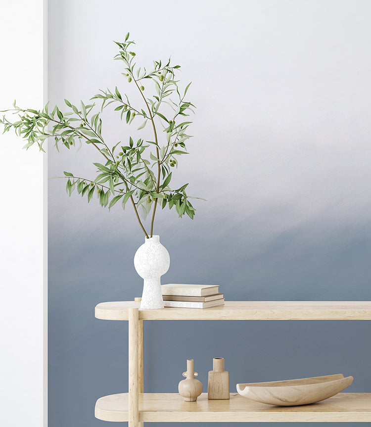 The room features the Delilah Waves, Watercolor Wallpaper in Blue, which transitions from white to calming shades of blue. Against this backdrop, a light wooden shelf holds a potted plant, books, and minimalist decor pieces. 