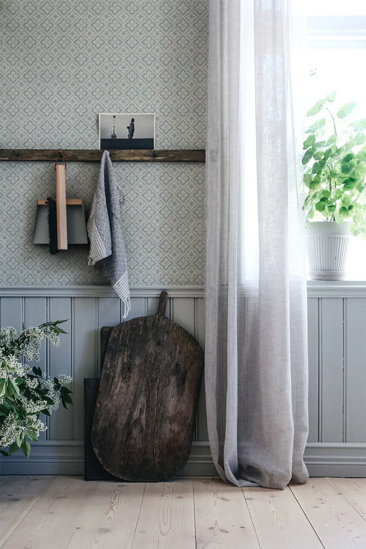 Edvin Rhombus Patterned Wallpaper in dusky blue featured on a wall of a room near near a window with white curtain and wooden equipment