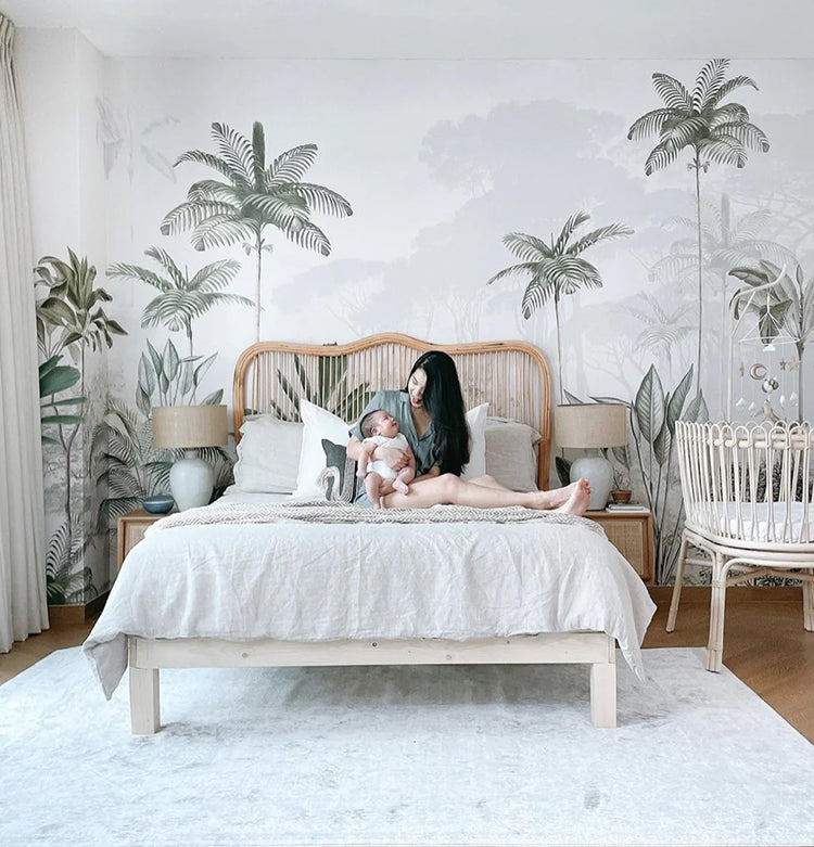 A serene bedroom with a cozy bed, wooden nightstands, and elegant lighting. A person is seen holding a baby. The room is adorned with an Elegant Rainforest, Tropical Mural Wallpaper in White.