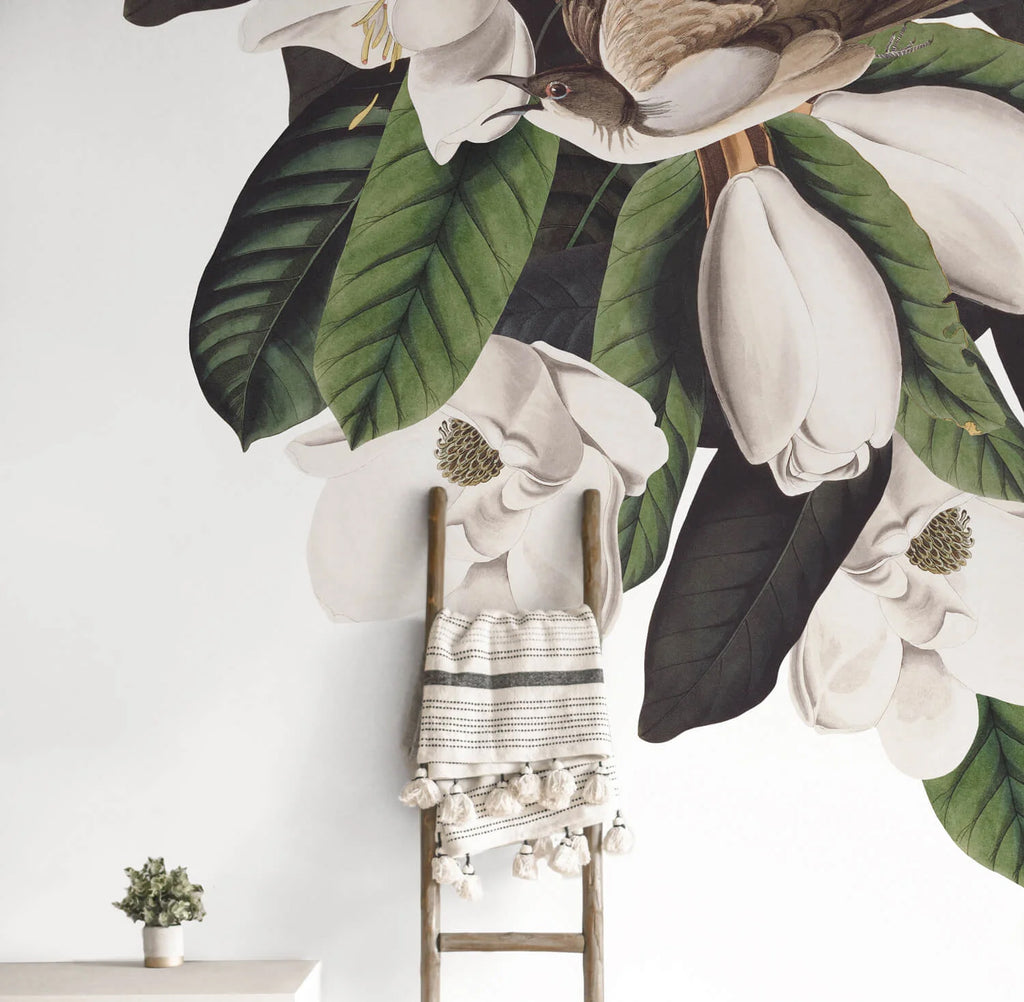 A lush mural Flora and Fauna, Mural Wallpaper . An old-fashioned wooden chair with a striped blanket is in front, with a small table to the left.