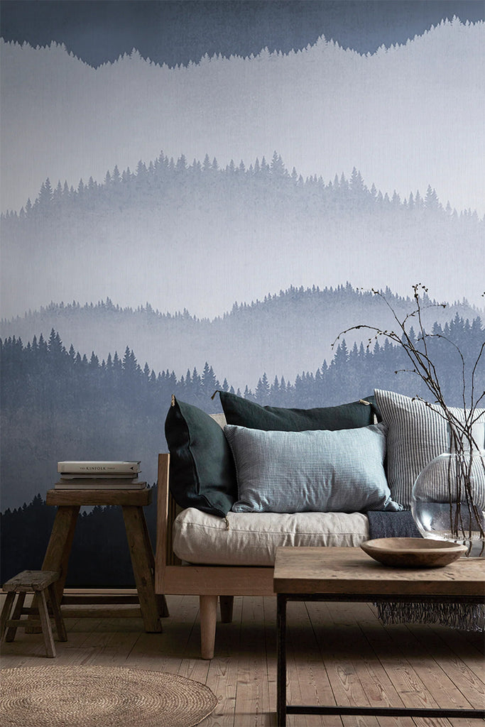 Forest Landscape, Ombre Mural Wallpaper in Dark Blue featured in a wall of a living area with wooden furnitures and flooring, with an assortment of pillows on top of a cushion. 