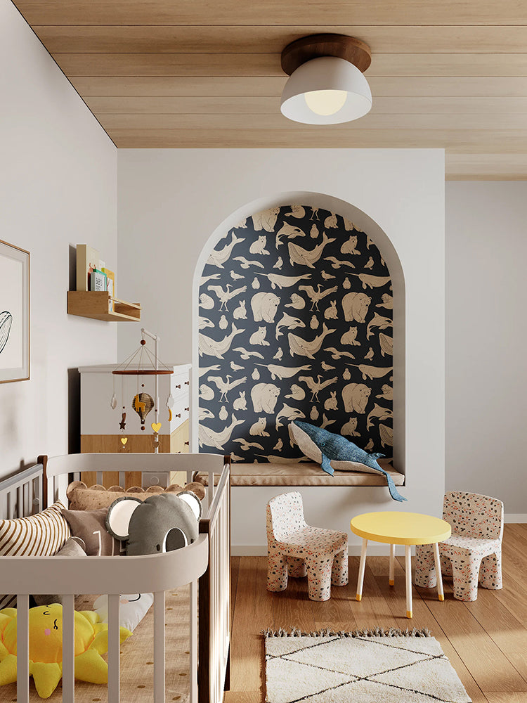 A modern nursery with wooden flooring, a white crib, matching dresser, and shelves filled with toys and books. A whimsical whale illustration adorns the wall. The room is accented by a round yellow table, and two small chairs with animal motifs. The alcove features the Frosty Friends, Animal Pattern Wallpaper in Blue.