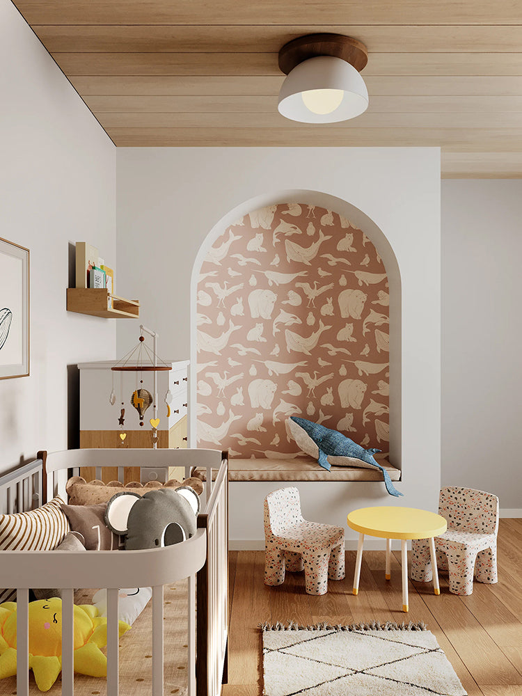 A modern nursery with wooden flooring, a white crib, matching dresser, and shelves filled with toys and books. A whimsical whale illustration adorns the wall. The room is accented by a round yellow table, and two small chairs with animal motifs. The alcove features the Frosty Friends, Animal Pattern Wallpaper in Dusty Pink.
