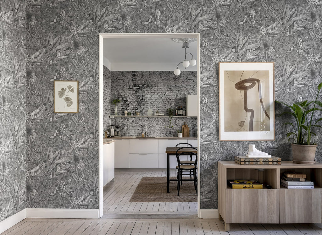 A stylish room with  Furada Floral & Toucan Pattern Wallpaper in Dark Grey, wooden floor, framed artwork, and a glimpse of a modern kitchen through an open doorway.