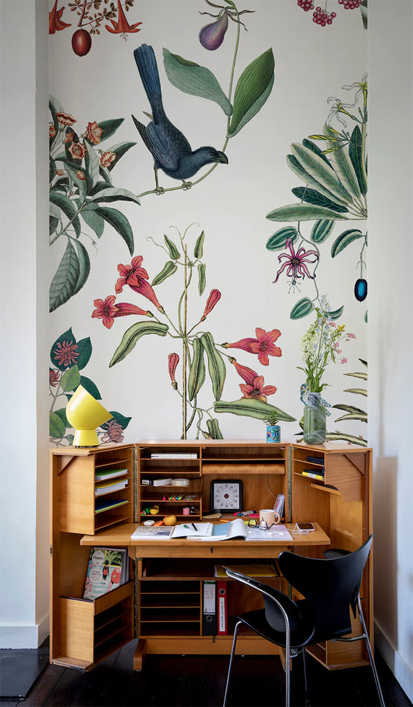A workspace with Garden Party, Mural Wallpaper  featuring colorful botanical illustrations and a bird. The wallpaper complements a wooden desk filled with books and stationery, paired with a black chair.