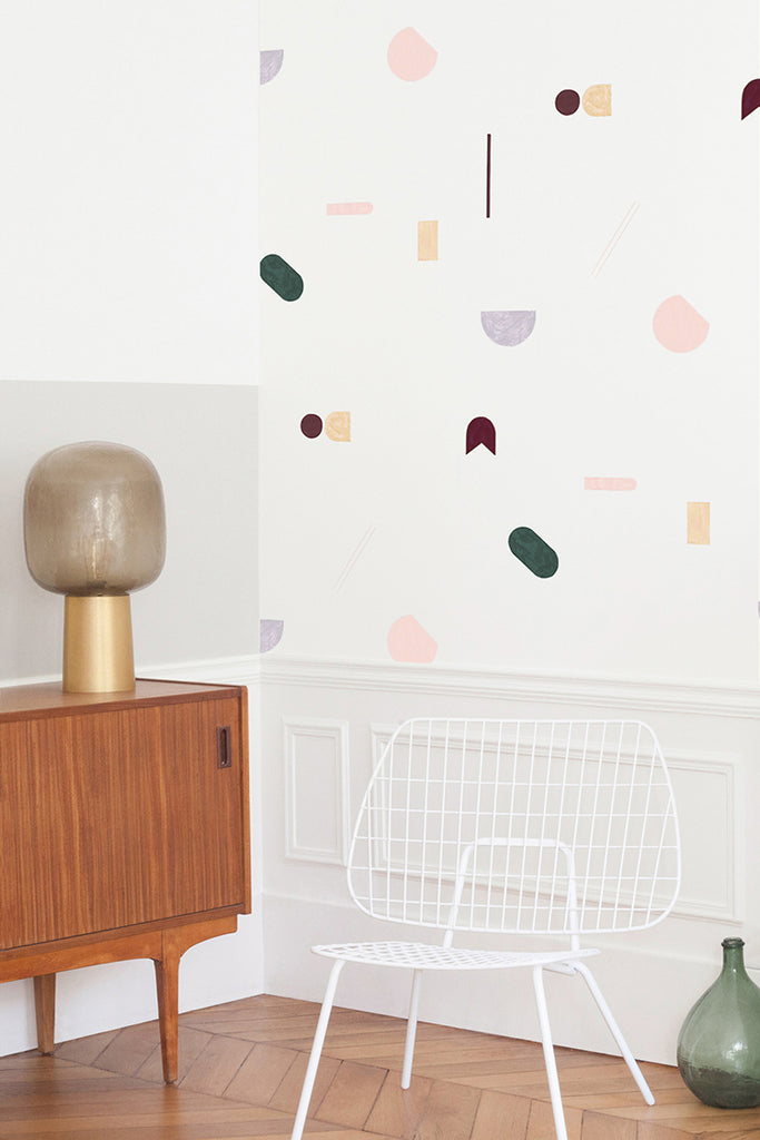 Geometric Canele, Wallpaper in Dark Green/Crimson featured on a wall of a room with a wooden sideboard that has a golden lamp on top and a chair made of white mesh wire