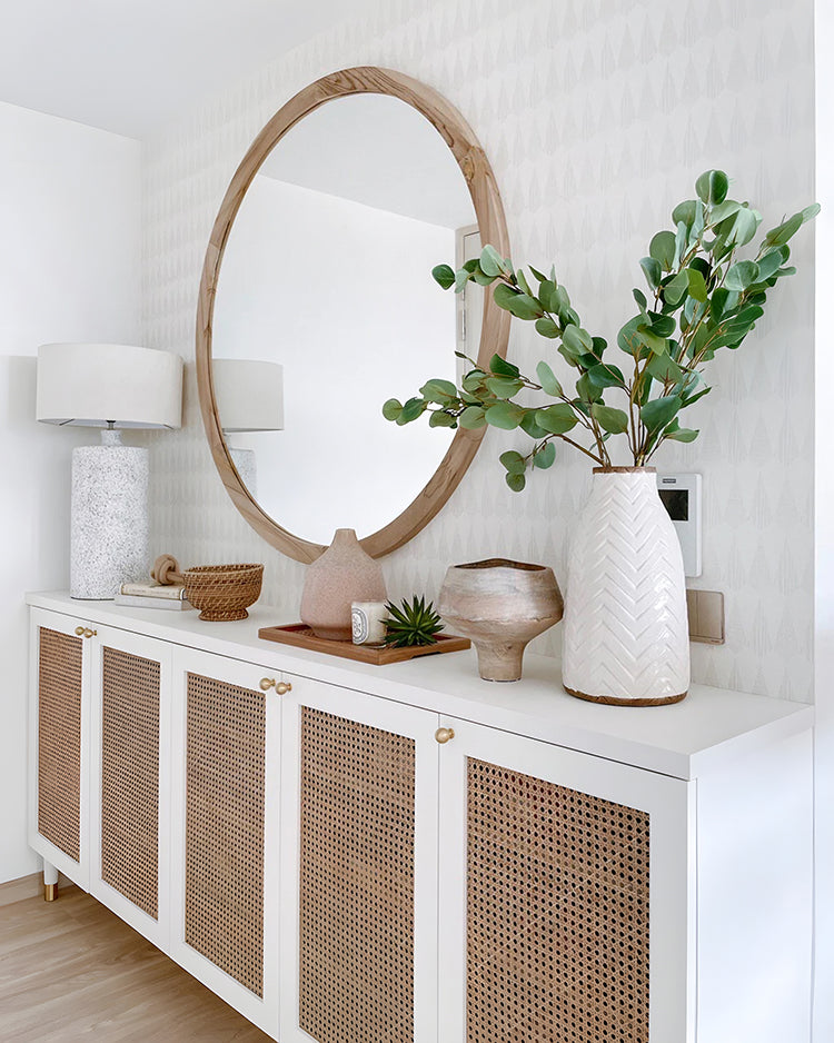 A well-lit room featuring a white cabinet with woven panels, adorned with a variety of decorative items. A large round mirror hangs on a wall covered with Geometric Panorama pattern wallpaper.