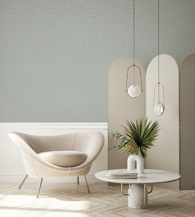 The room features a modern beige armchair and a round marble table. The Gold Metallic Confetti Speckles, Pattern Wallpaper Blue adds a touch of luxury, with pendant lights hanging above. A potted plant provides a fresh contrast to the metallic accents, creating an elegant and inviting space.