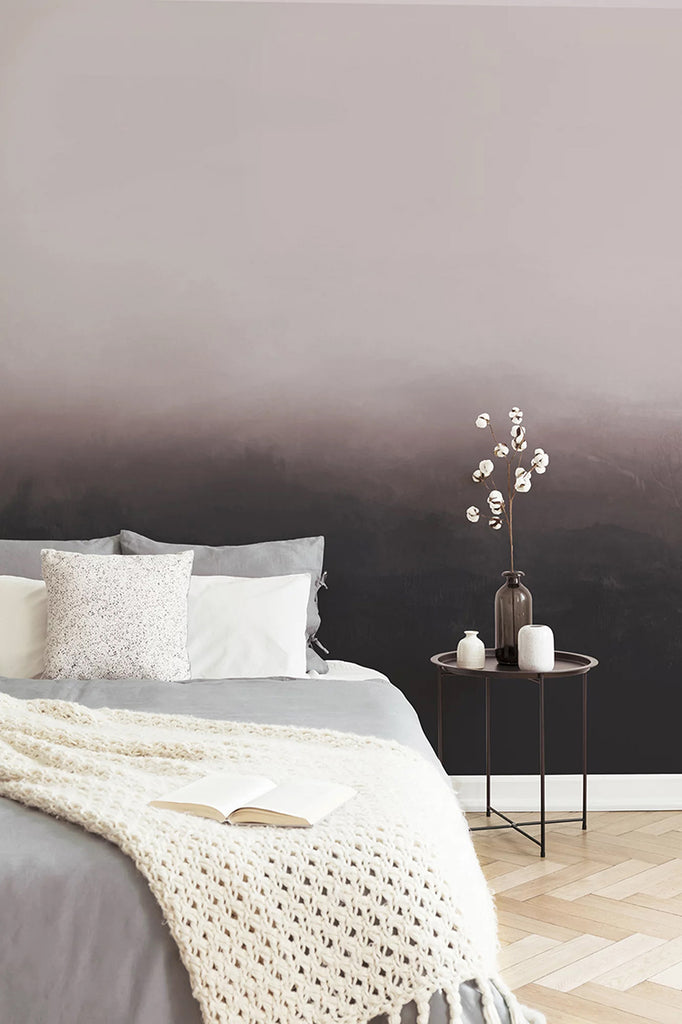 Greg Horizon Ombre, Wallpaper in Burgundy close featured on a wall of a bedroom and a bed with grey sheets and multiple pillows, beside it is a side table with a vase and ceramic containers 