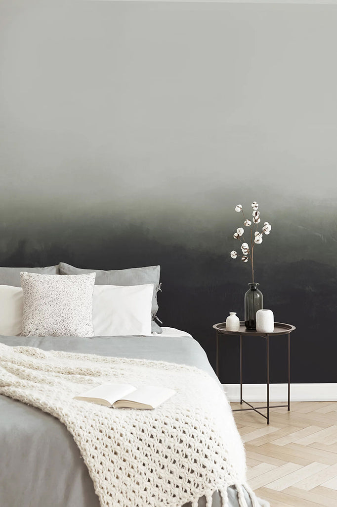Greg Horizon Ombre, Wallpaper in Dark Green close featured on a wall of a bedroom and a bed with grey sheets and multiple pillows. Thereis a side table with a vase and ceramic containers 