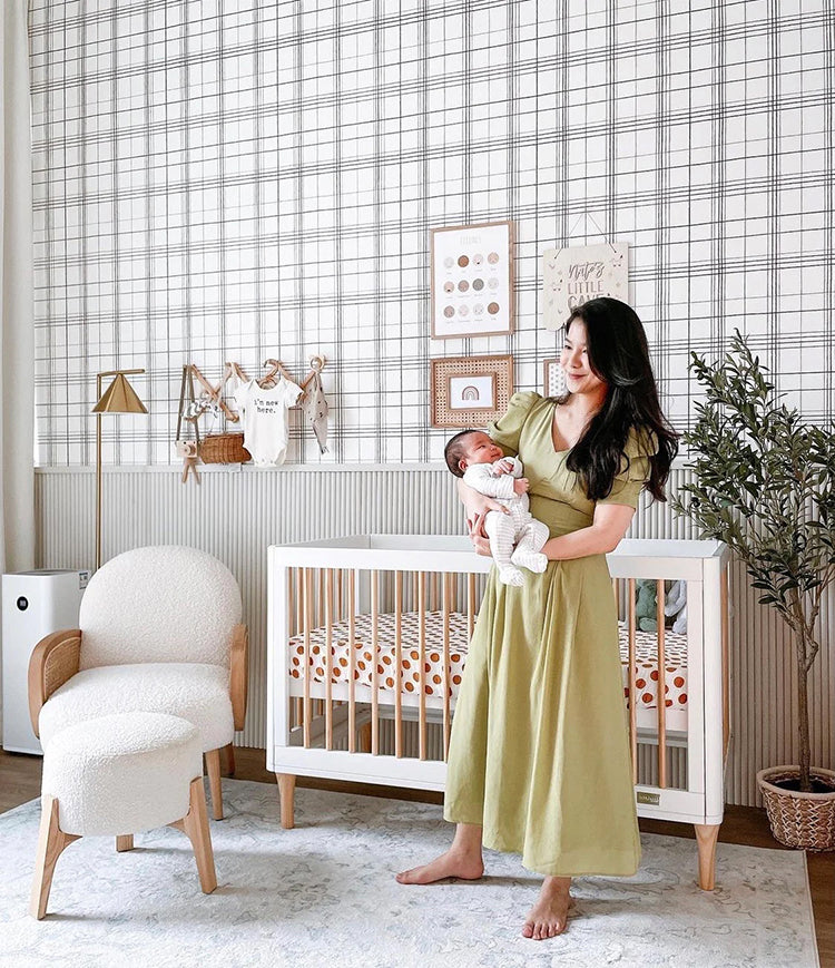 A nursery with Hand-Drawn Plaids, Pattern Wallpaper in Black, featuring a crib, a chair, wooden decor, and a person holding a baby in soft lighting.