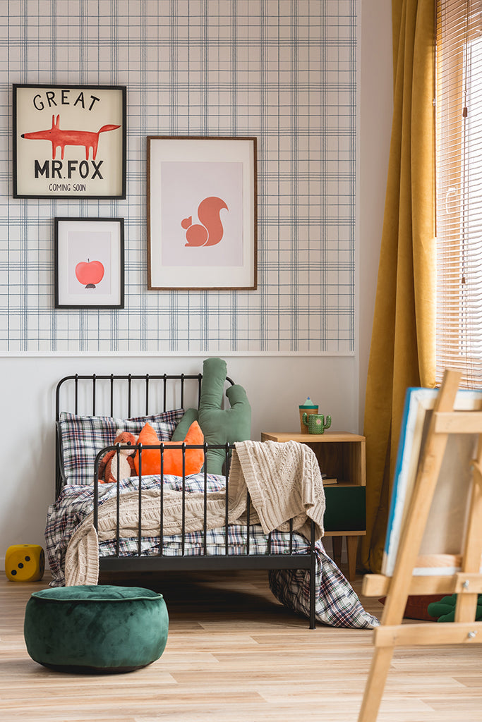 A cozy, well-lit room with a metal bed adorned with plaid bedding and green cushions. Framed artworks hang above the bed against a Hand-Drawn Plaids, Pattern Wallpaper in Black . A wooden side table with a green lamp and a dark green ottoman add to the room’s charm.