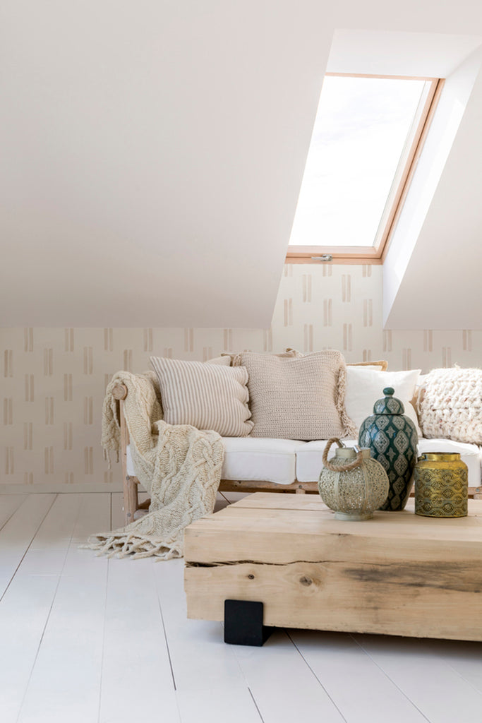 An attic room with a skylight, featuring a comfortable sofa with textured cushions, a rustic wooden table, and decorative vases. The room is illuminated by natural light, creating a cozy ambiance. The walls are adorned with Hand-drawn Duo Stripes wallpaper in Beige.