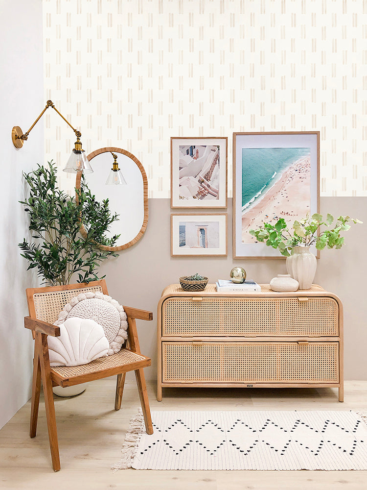 A minimalist room with a wooden chair, woven cabinet, framed art, plants, and a mirror. The room is lit by a brass wall lamp, and the floor is adorned with a geometric rug. The backdrop is a Hand-drawn Duo Stripes pattern wallpaper in Beige.