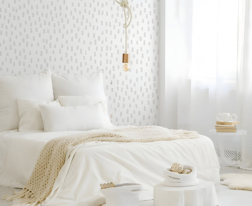 Sunlit bedroom with white bedding and cream throw. Wooden stool with books and plant. Woven baskets on floor. Hand Drawn Ovals decals on wall.