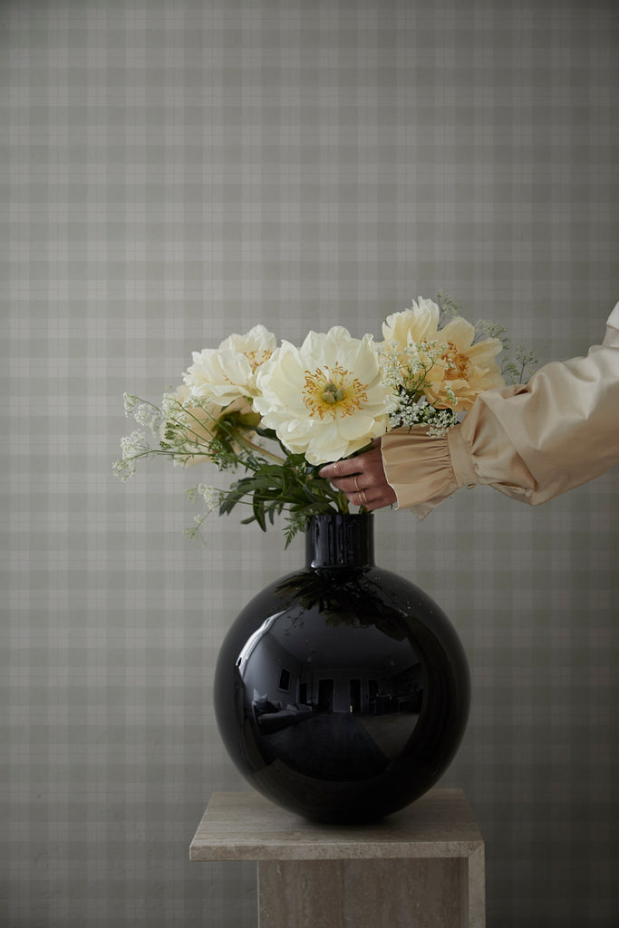 Herman, Plaid Pattern Wallpaper in grey featured on a wall of room with a a stool with a black flower vase and a yellow flower