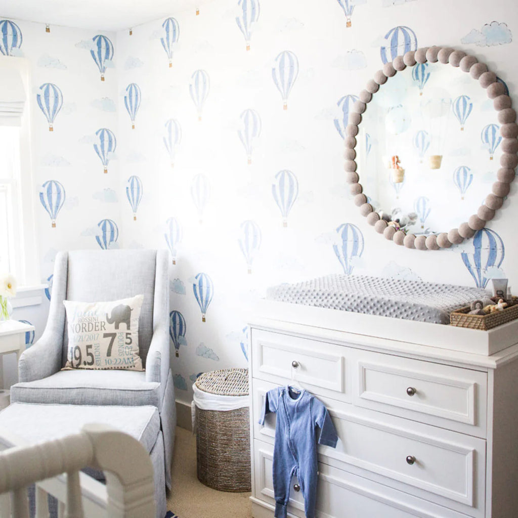 A well-lit nursery room with a Hot Air Balloon, Pattern Wallpaper . A comfy grey armchair, a white dresser with a changing pad, and a woven basket are visible.