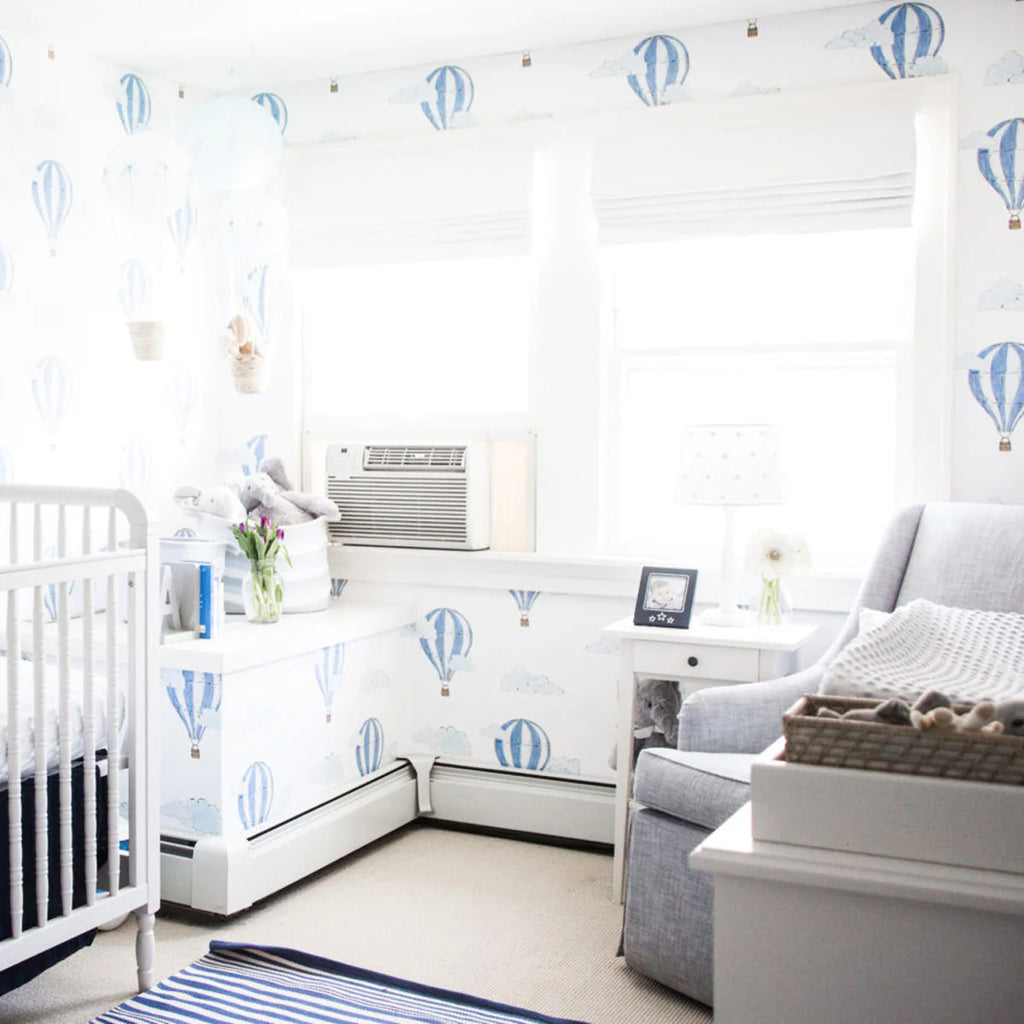 A bright and airy room with Hot Air Balloon Patterned Wallpaper. Features include a white crib, a comfy grey chair with a white throw blanket, and a small white table with decorative items.