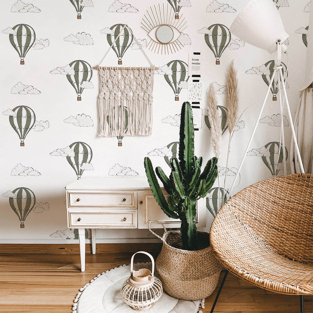 A room with a green Hot Air Balloon pattern wallpaper, a white dresser, a large cactus in a basket, a rattan chair, and decorative items creating a warm, inviting space.