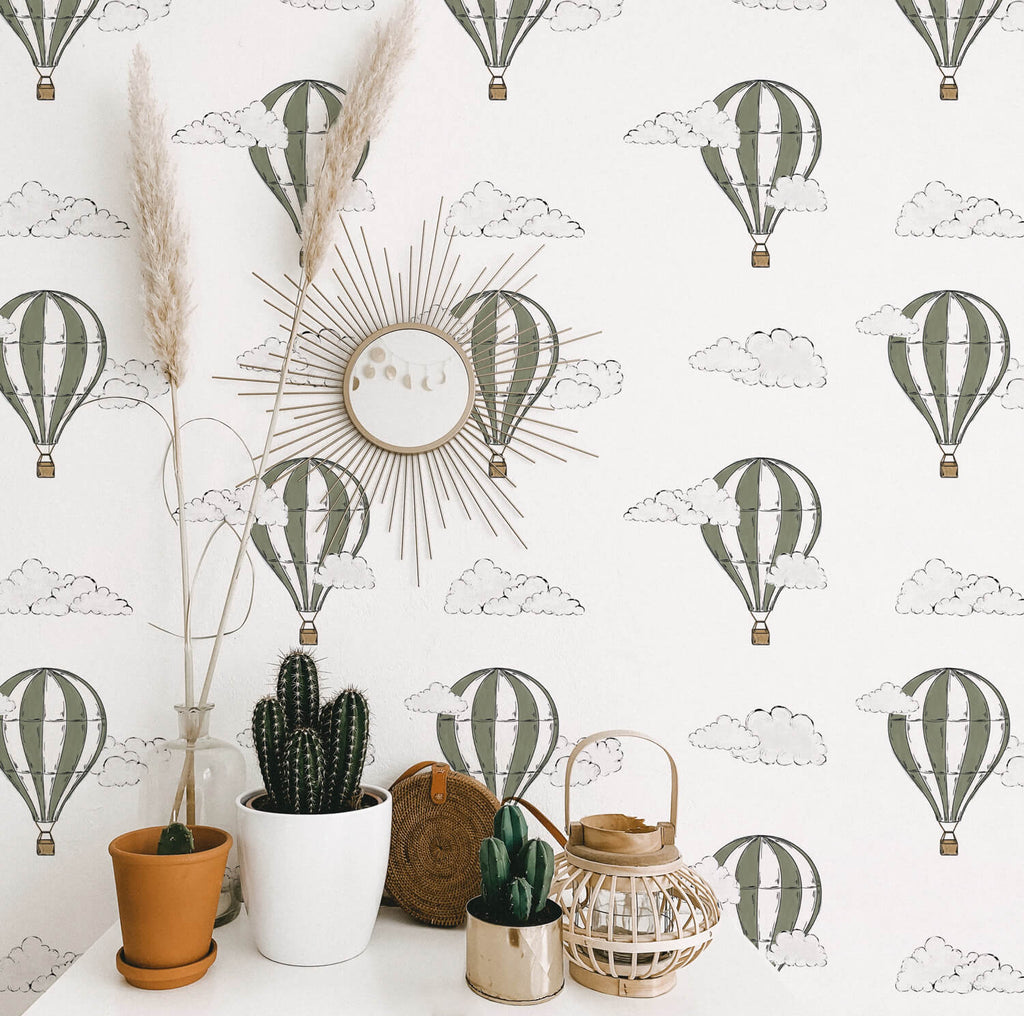 A wall featuring  Hot Air Balloon, Pattern Wallpaper in Green. A sunburst mirror is centrally placed on the wall. Various potted plants including a tall plant with feathery plumes, a cactus, and smaller green plants are in front of the wall. A woven basket adds texture to the scene.
