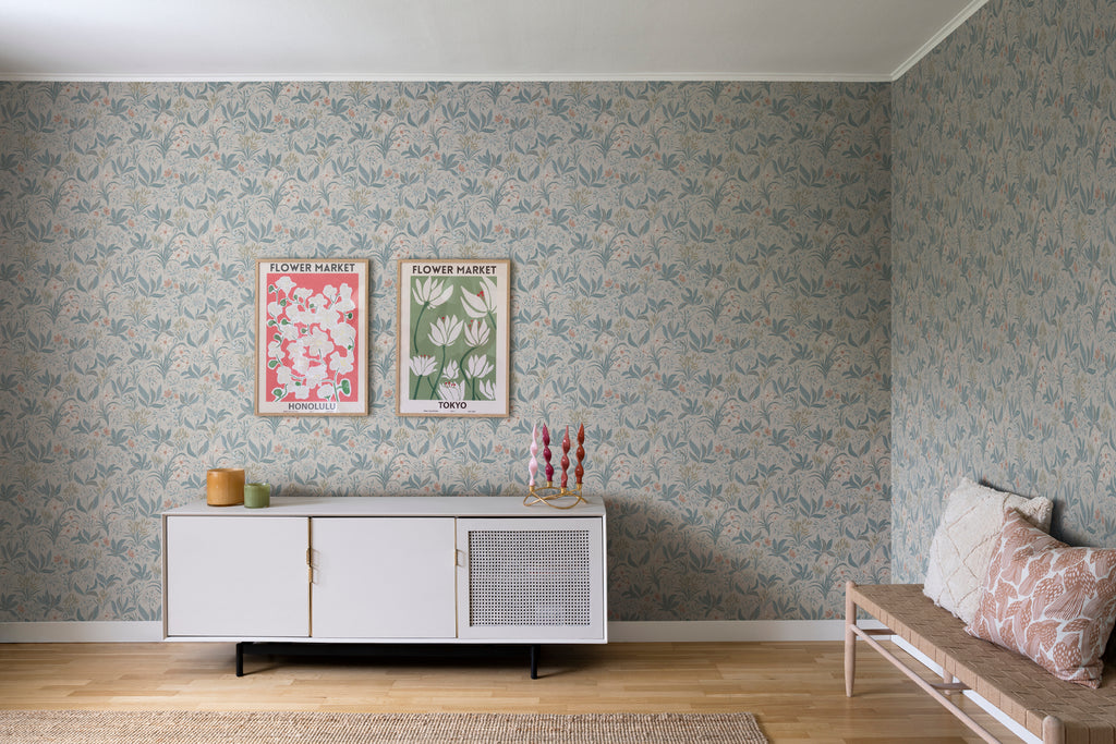 Huset i Solen, Floral Pattern Wallpaper in sand featured in a room with a white cabinet that has candles and ceramic mugs on top of it, also seen is a rattan long chair with two different designs of pillows