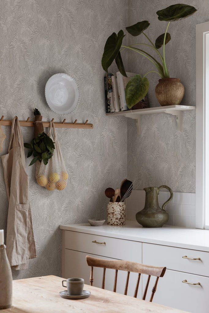Idun, Nature Pattern Wallpaper in grey featured on a wall of a kitchen with white counter with ceramics non top, a wooden dining table, and an apron and groceries hanging on the wall. 