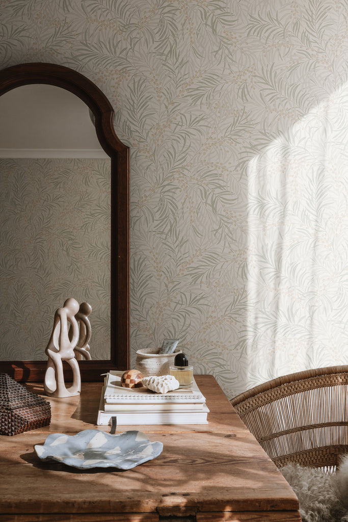 Idun, Nature Pattern Wallpaper in Sand featured on a wall of a room with wooden table with books, ceramics, and mirror on top, and a Rattan Chair. 