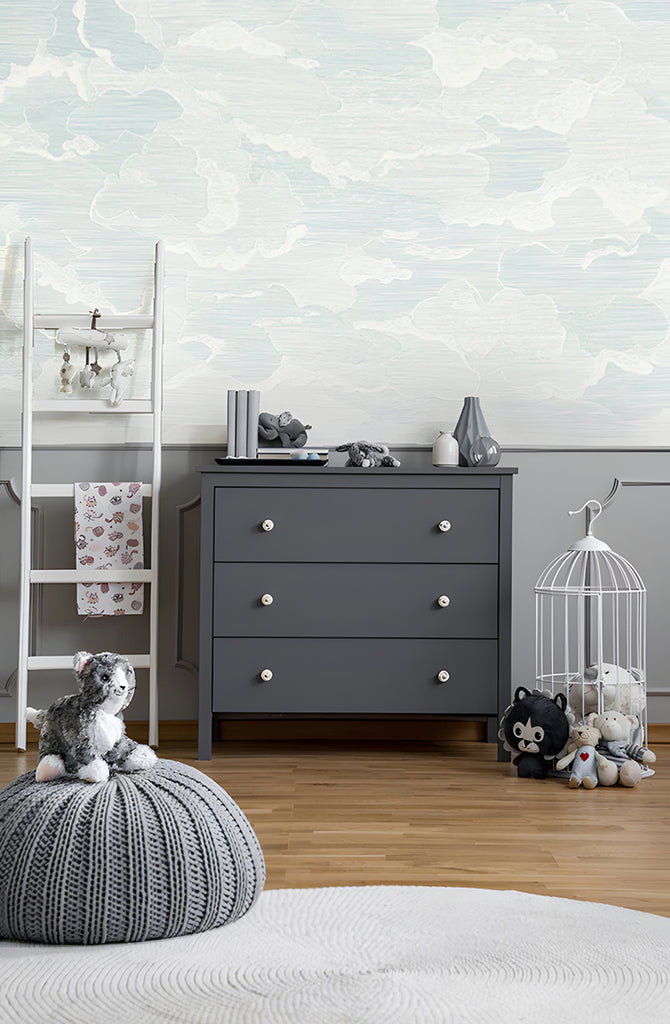 Illustrated Clouds, Mural Wallpaper in Blue, featured on a room wall, that brings  whimsical and serene atmosphere . This room is adorned with a grey cabinet, on top of which ceramics are displayed. Plush toys are scattered around the room, adding a playful touch to the space. The blue mural wallpaper with its illustrated clouds brings a whimsical and serene atmosphere to the room.