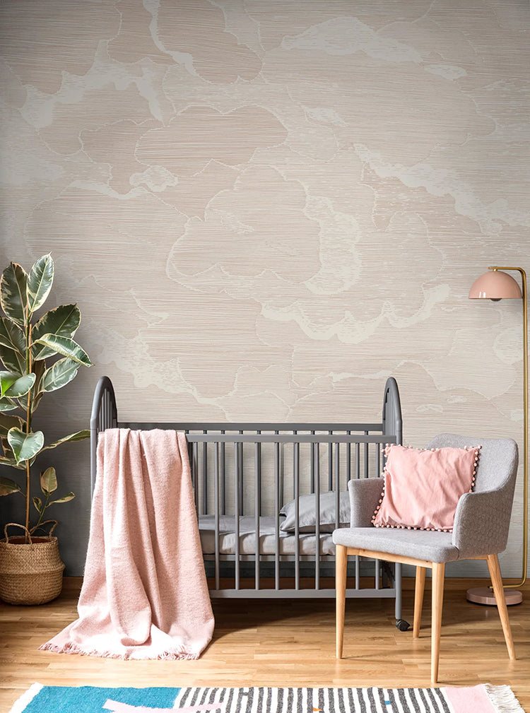 Illustrated Clouds, Mural Wallpaper in Grey sets a calming tone in a child’s nursery room. The room is furnished with a grey crib, adorned with a pink blanket, and a grey chair that is made comfortable with a pink pillow