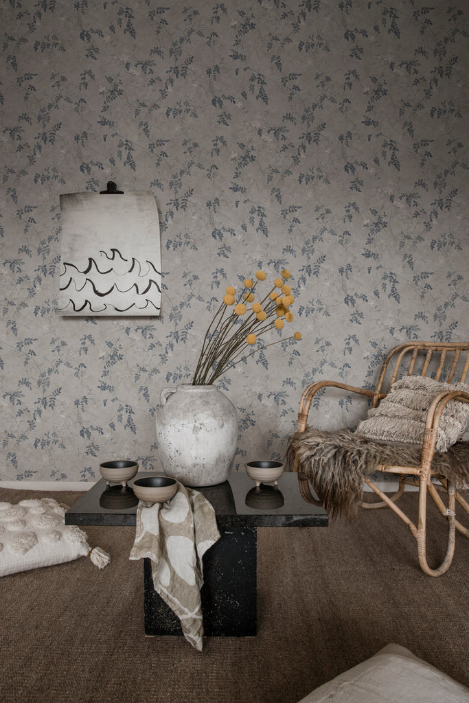 Irene, Floral Pattern Wallpaper in Blue featured on a wall of a room with a rattan chair and black coffee table, and on top is a white vase with flowers