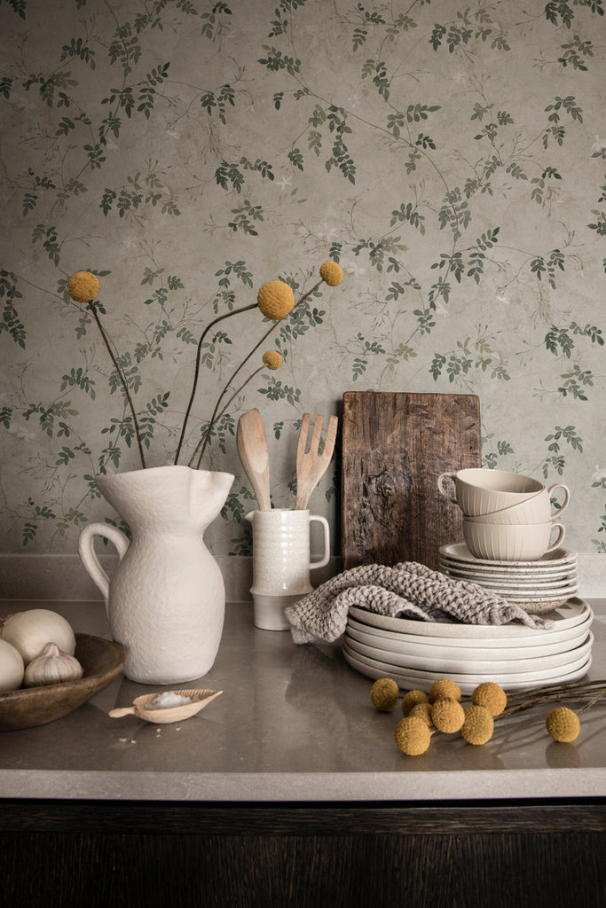 Irene, Floral Pattern Wallpaper in Green featured on a wall of a kitchen with a table with stacked plates and several kitchenwares on top