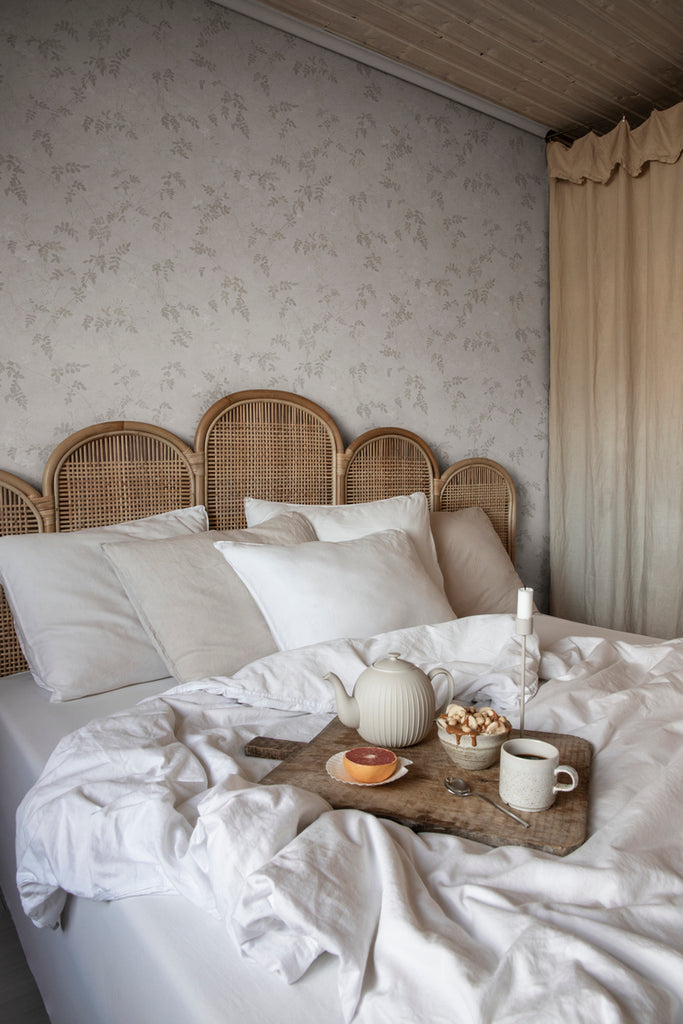 Irene, Floral Pattern Wallpaper in Grey featured on a wall of a bedroom with a bed that has a rattan frame and white pillows and sheets
