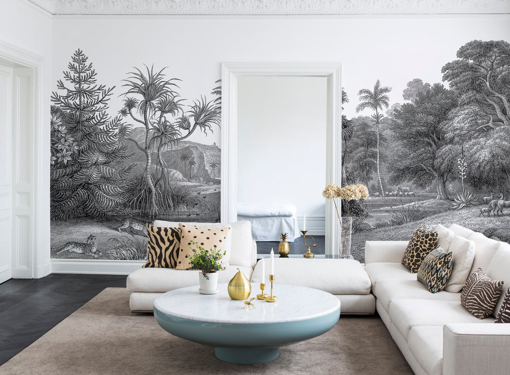 Jungle Land, Mural Wallpaper in Stratos Grey featured on a wall of a living area with grey sofa and round table with plants on it