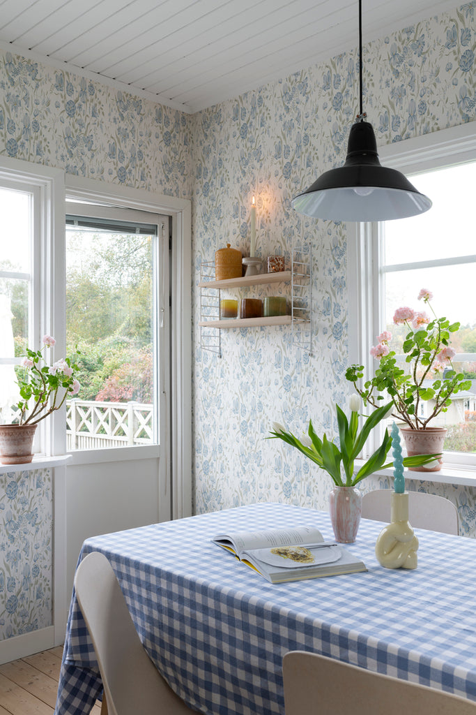Karins Bukett, Floral Pattern Wallpaper in White featured on a wall of a dining area with a dining table that has a blue checkered tablecloth with books, vase, and candle, along with a pendant light above.