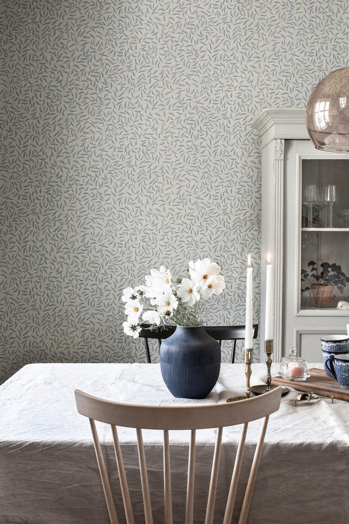 Karolina Foliage, Pattern Wallpaper in dark blue featured on a wall of a dining area with a wooden chair and a dining table with white table cloth 