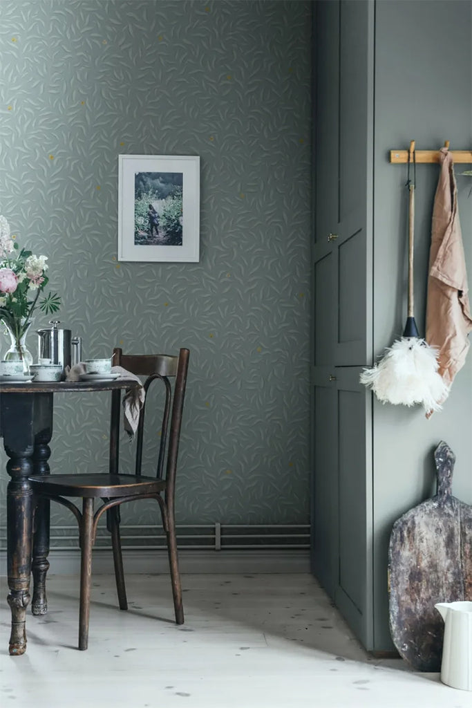 Karolina Foliage, Pattern Wallpaper in green, featured on a wall of a room with a vintage wooden table and chair.