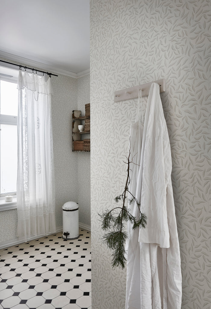 Karolina Foliage, Pattern Wallpaper in light grey featured on a wall of a room with clothes hanging on a wall 
