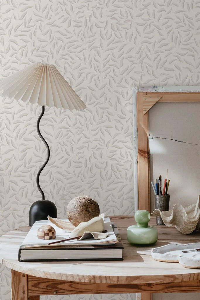 Karolina Foliage, Pattern Wallpaper in light grey featured on a wall of a room against a wooden round table with lamp, books and furnitures 