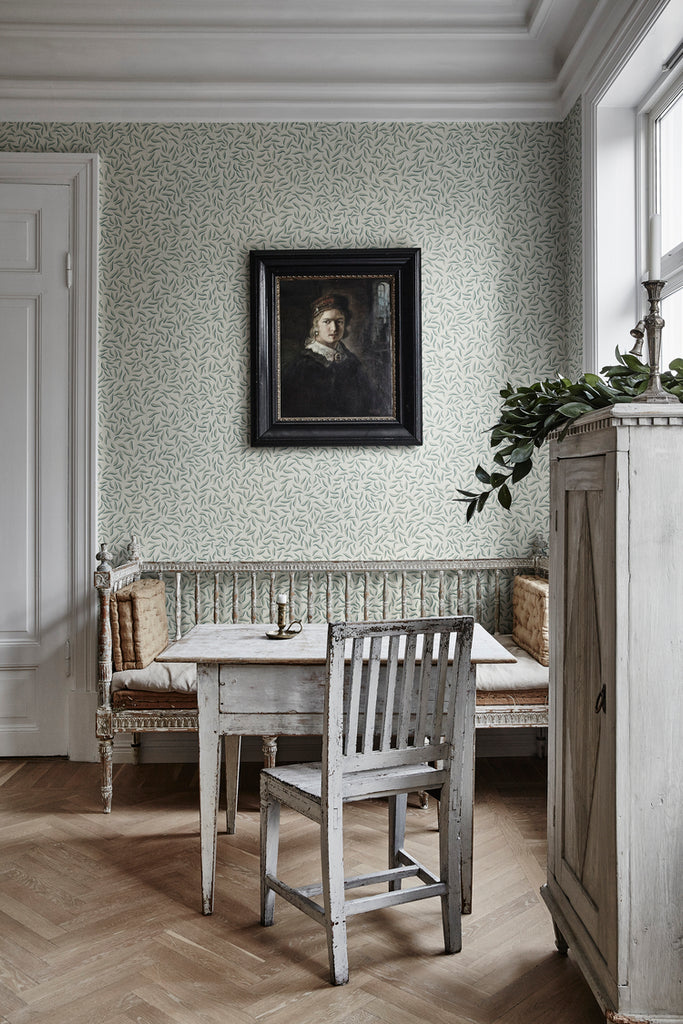 Karolina Foliage, Pattern Wallpaper in sage featured on a wall of a room with wooden furniture and wooden flooring